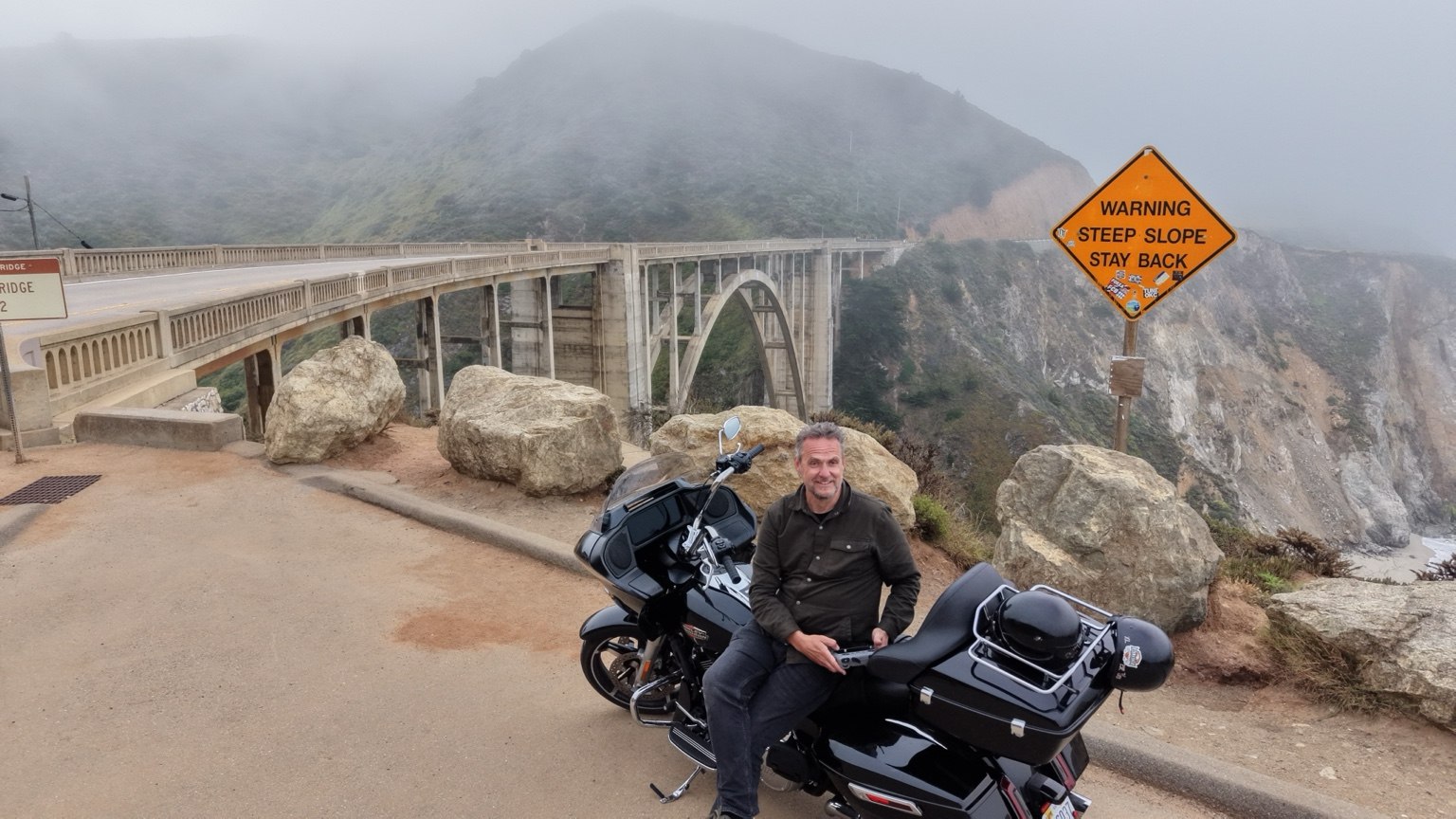 A motorcycle stop beside a coastal arch bridge in the mist